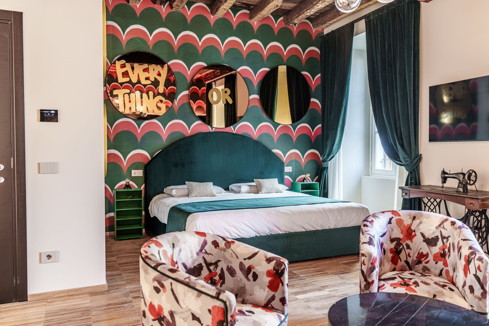 A bedroom with a teal headboard, a bed with white linens, a green nightstand, a floral armchair, and a vintage sewing machine.