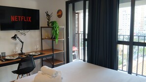 Habitación de hotel con cama, escritorio con una laptop, televisor montado en la pared, planta en una estantería y vista de la ciudad a través de amplias ventanas.