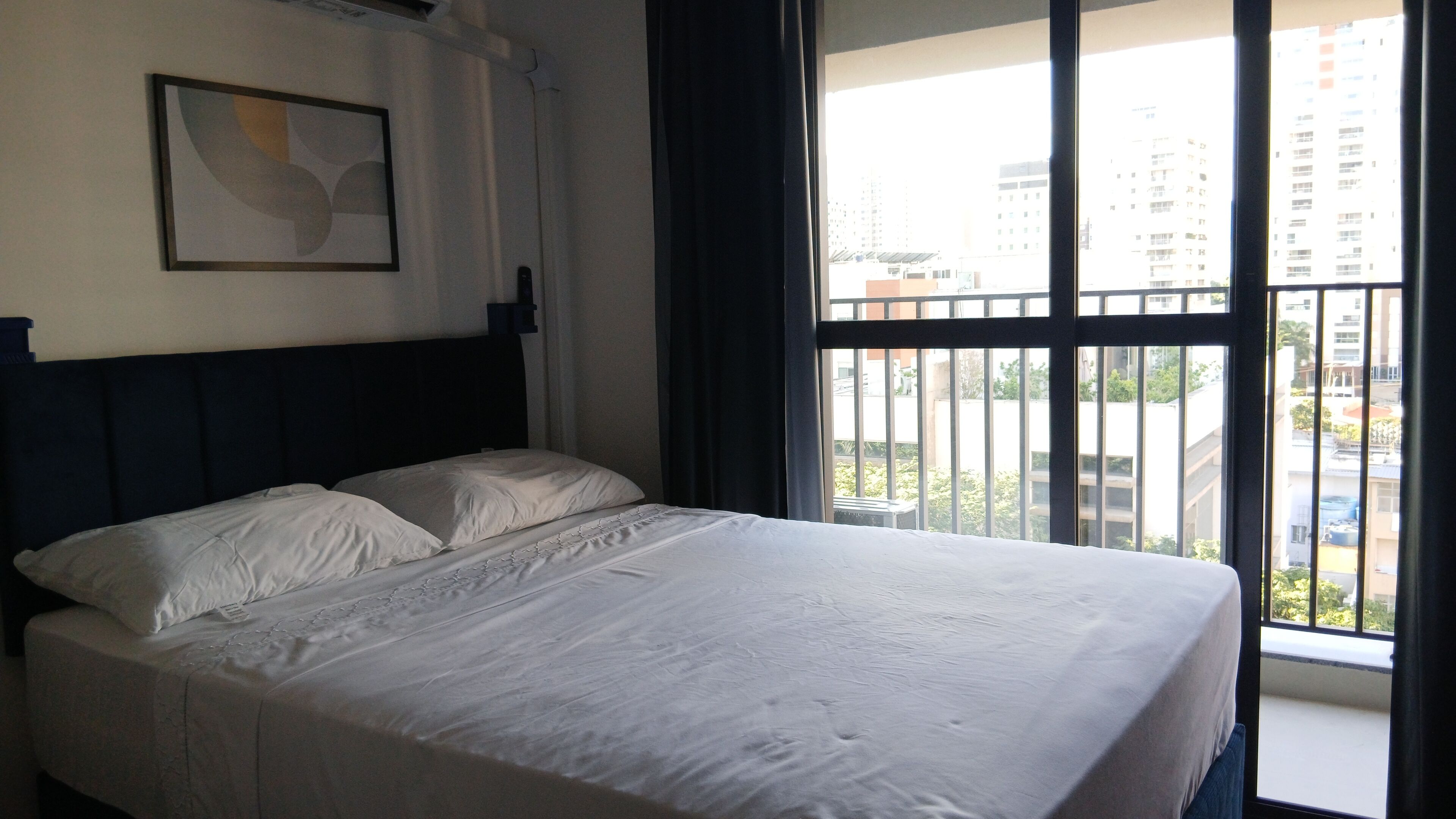 Una habitación de hotel con una cama grande, cabecero, ventana con vista a edificios y un cuadro en la pared.