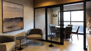 Una habitación de hotel moderna con un ventanal grande, un área de comedor con mesa y sillas, un sofá y un cuadro en la pared.