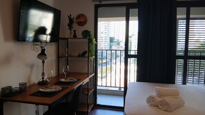 A hotel room with a bed, a desk with a lamp, a TV, a balcony with a view of buildings, and a window with curtains. - Vista incrível com AC e academia (São Paulo)
