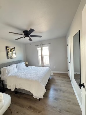 A bedroom with a bed, a ceiling fan, a window with curtains, a bedside table with a lamp, and a large mirror on the wall.