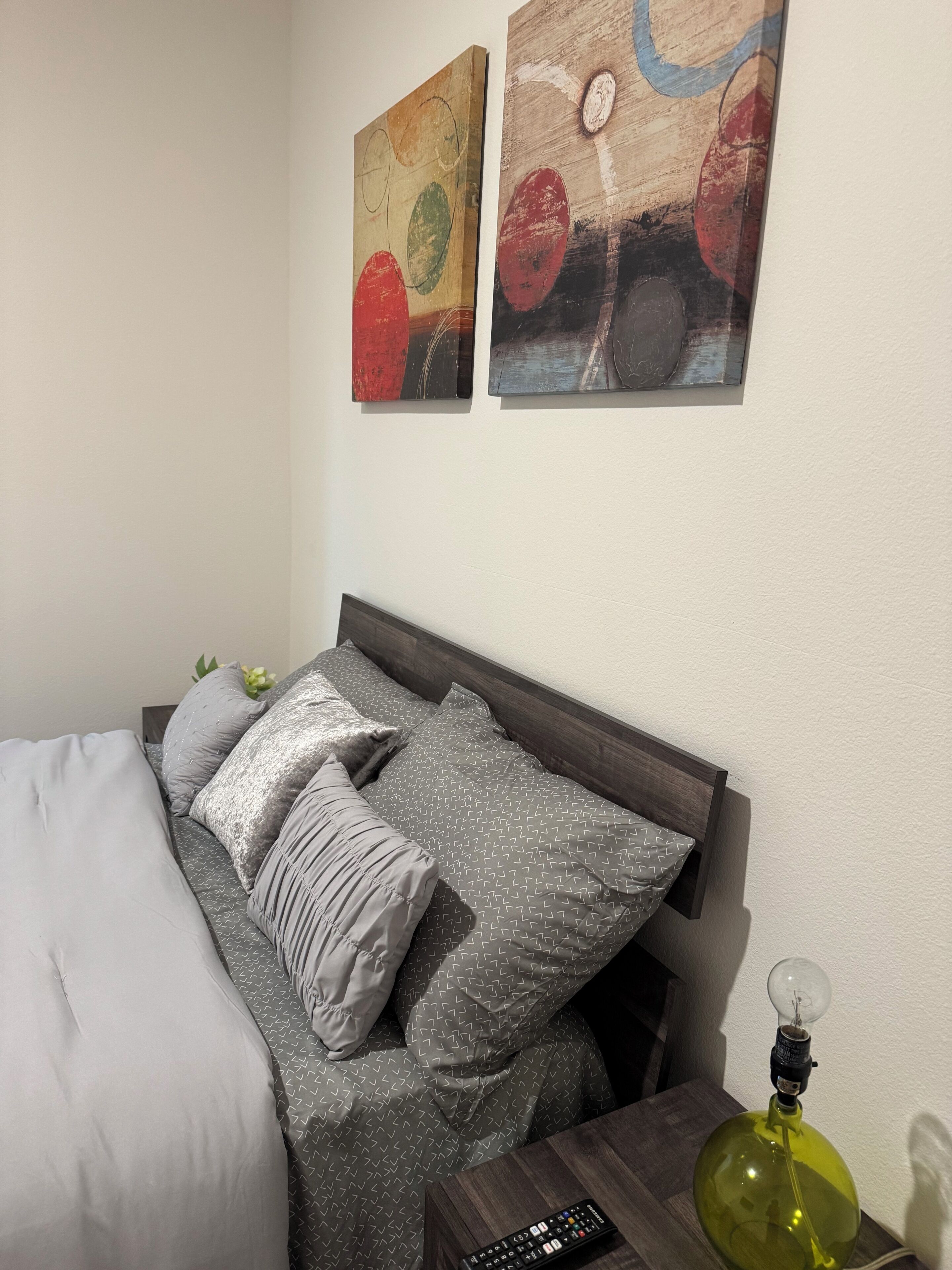 Een bed met grijs beddengoed met een patroon, een afstandsbediening en een groene lamp op een nachtkastje.