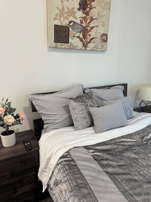 Ein ordentlich bezogenes Bett mit einer grau-weißen Decke, einem dunklen Holzkopfschild und einem kleinen Topf mit rosa Blüten auf einem Nachttisch.