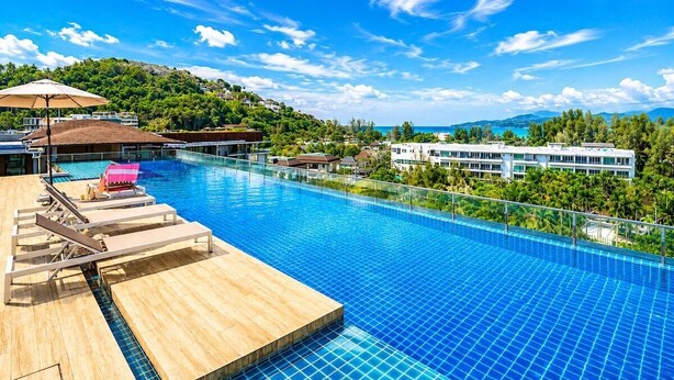 Ein Schwimmbecken mit Liegestühlen, einer Holzterrasse und Blick auf einen Hügel mit Gebäuden.