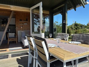 Un espace repas extérieur avec une table en bois, des chaises blanches et une porte vitrée donnant sur une cuisine.