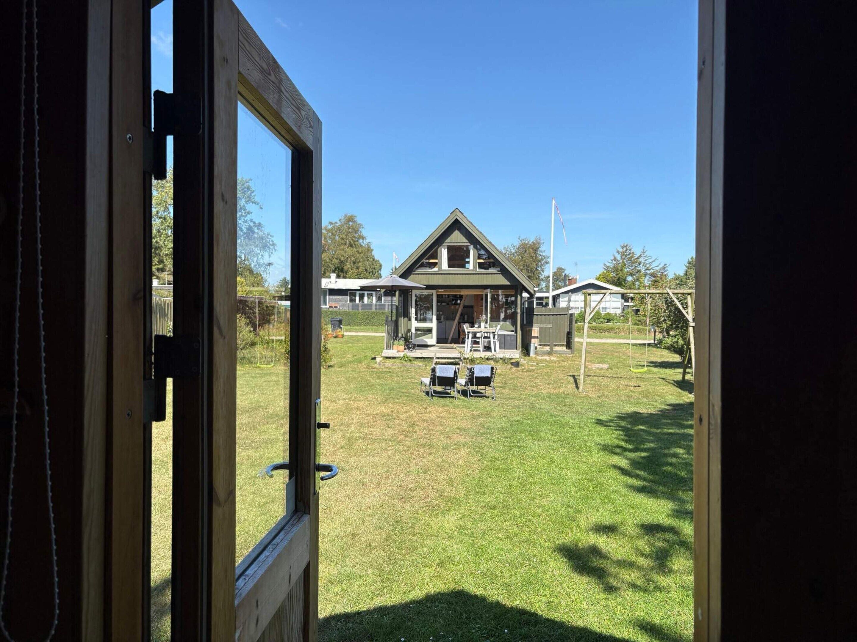 Vista a través de una puerta de madera que muestra una casa con césped, mobiliario exterior y un columpio.