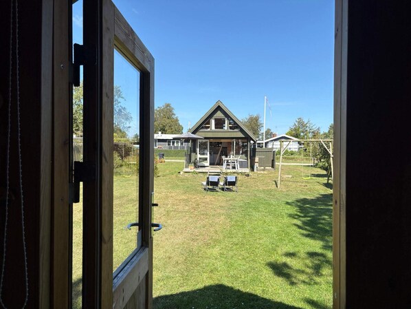 On aperçoit, à travers une porte en bois, une maison avec une pelouse, un espace salon extérieur et une balançoire.
