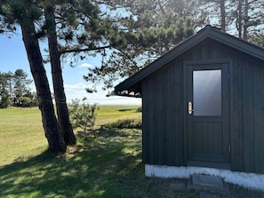 Un petit abri en bois sombre, avec une base blanche, entouré d’herbe et d’arbres.