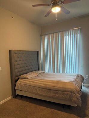 A bedroom with a tufted headboard, a bed with striped bedding, a ceiling fan, and a window with vertical blinds.