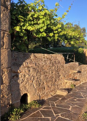 A stone wall with a small opening, a stone-paved path, and green foliage. - Renovated farmhouse dating from 1821 (SAINTE HELENE)
