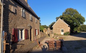 A stone building with a paved entrance, a small outdoor seating area with wooden furniture, and a clear blue sky. - Renovated farmhouse dating from 1821 (SAINTE HELENE)
