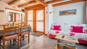Uma sala de estar aconchegante com vigas de madeira, uma mesa de jantar com cadeiras, um sofá com estofamento floral e uma pequena mesa com livros e um controle remoto.