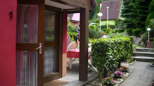 Um edifício com fachada vermelha, uma porta com janela e um pequeno jardim com plantas e uma cadeira.