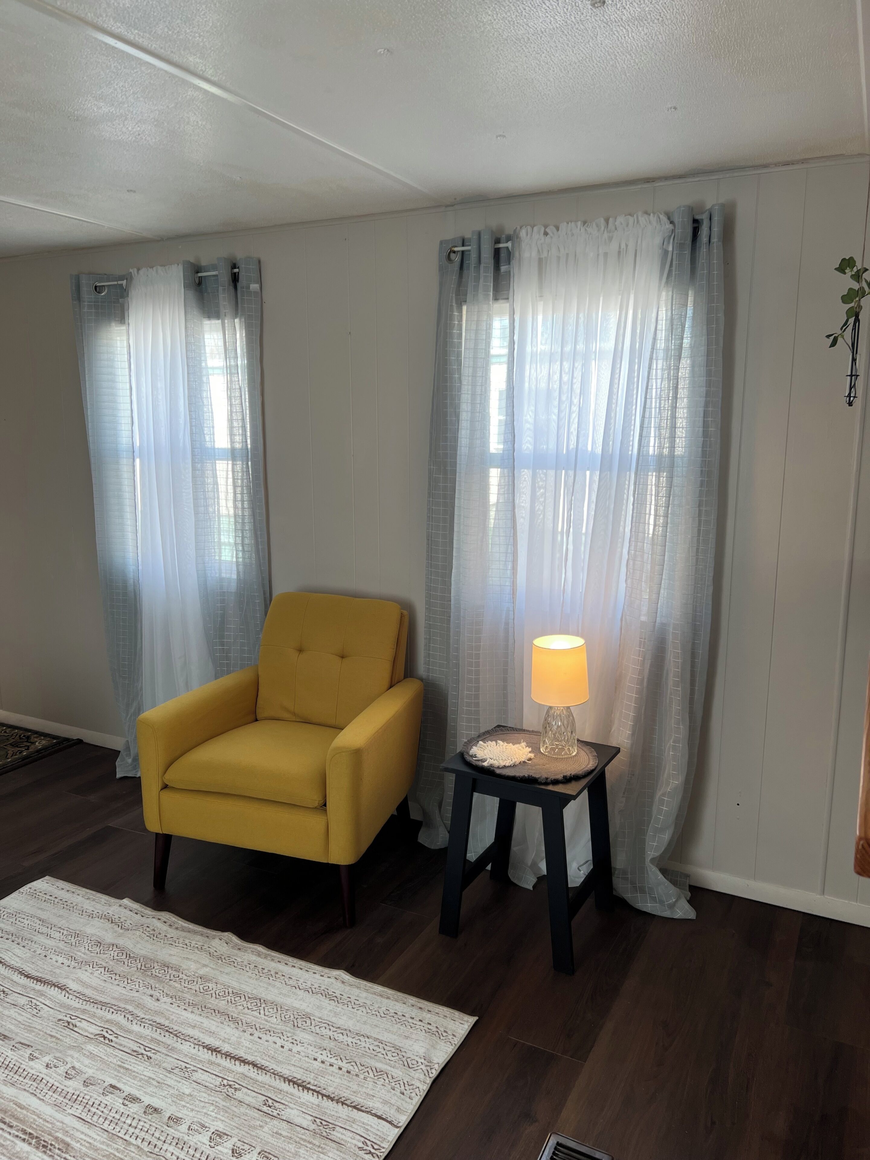 A room with a yellow armchair, a table with a lamp, and sheer curtains.