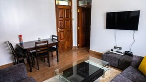 Ett vardagsrum med ett soffbord i glas, grå soffor, ett matbord i trä med stolar, en platt-TV som är monterad på väggen och en dörr som leder till ett annat rum.