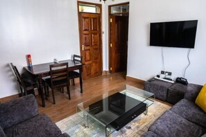 A living room with a glass coffee table, grey sofas, a wooden dining table with chairs, a flat-screen TV mounted on the wall, and a door leading to another room. - Karen Brooks View Palace- 2 Bedroom (Nairobi)