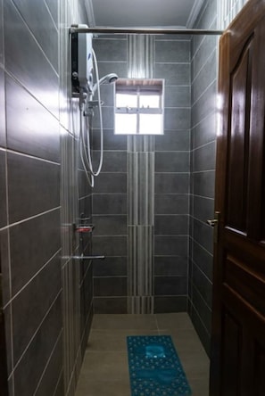 A narrow shower stall with dark tiles, a handheld showerhead, a small window, and a wooden door.