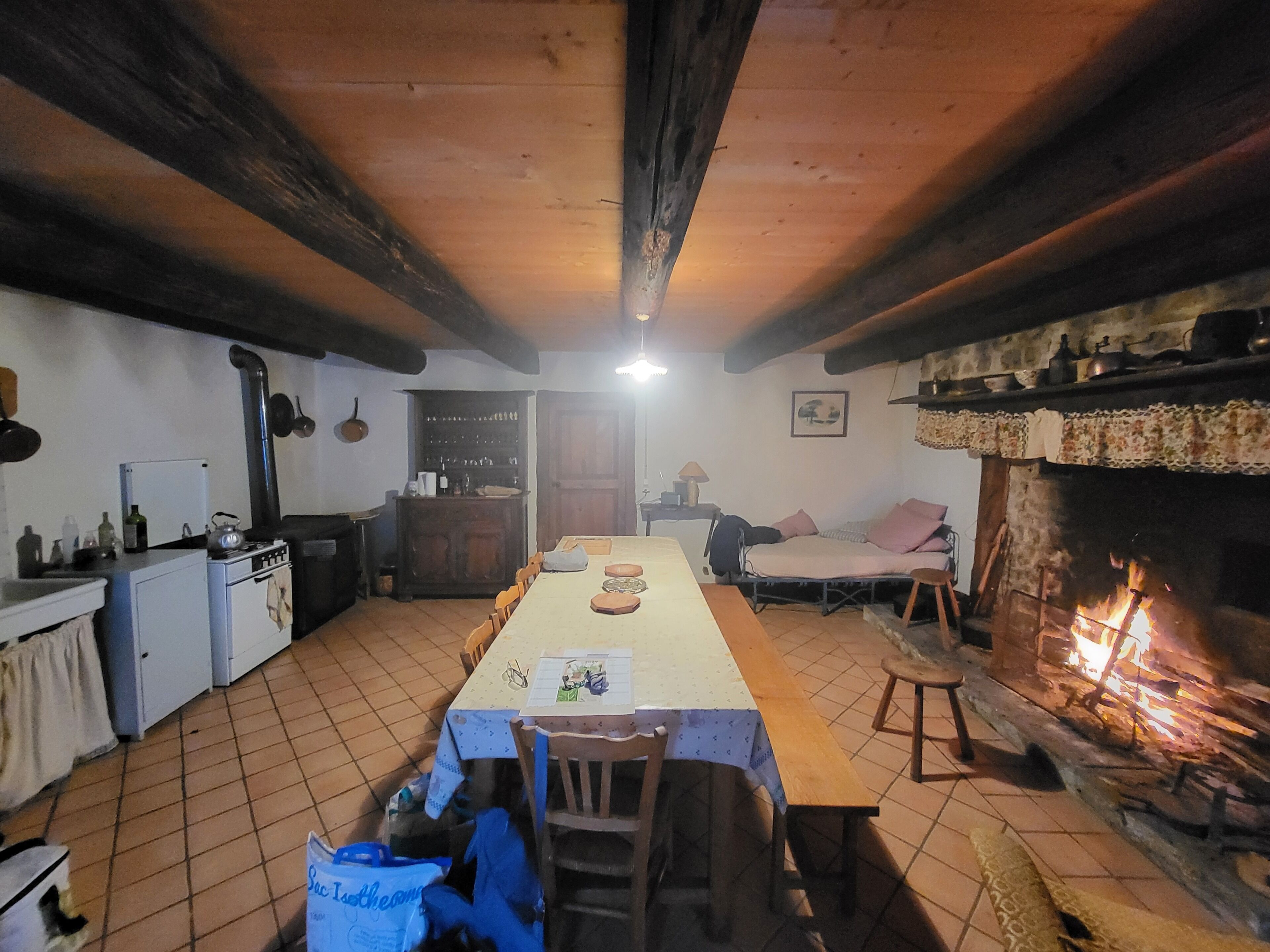 Una cocina rústica con suelo de baldosas, vigas de madera, una mesa de comedor con sillas, una chimenea y un refrigerador pequeño.