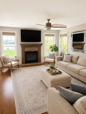 A living room with a fireplace, a ceiling fan, and a large window.