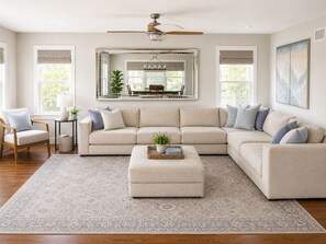 Ein Wohnzimmer mit einem Ecksofa, einem kleinen Tisch mit Pflanze, einem Deckenventilator und einem großen Spiegel über einem Esstisch.