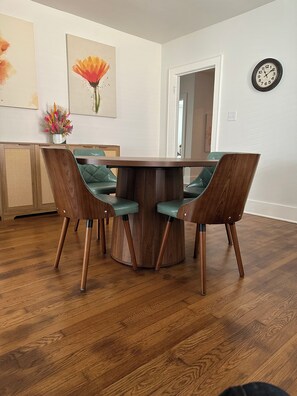 A dining area with a wooden table and four chairs, a clock on the wall, and artwork above the table. - Charming Vintage Coral House in a quiet neighborhood near downtown (Kerrville)