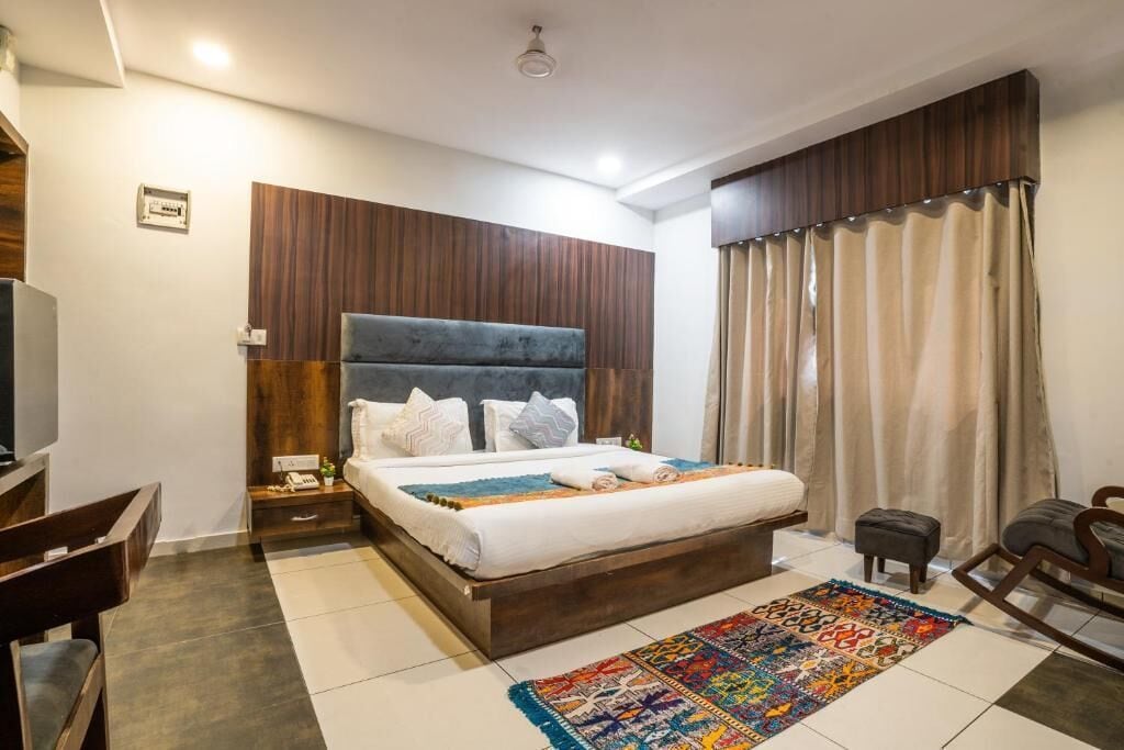 Quarto de hotel com uma cama, escrivaninha, cadeira, mesinha e janela com cortinas.