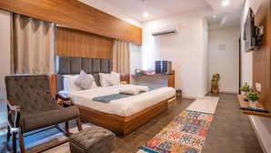 Un dormitorio moderno con cabecera de madera, una cama grande, un sillón tapizado en gris, un pequeño reposapiés y una alfombra colorida.