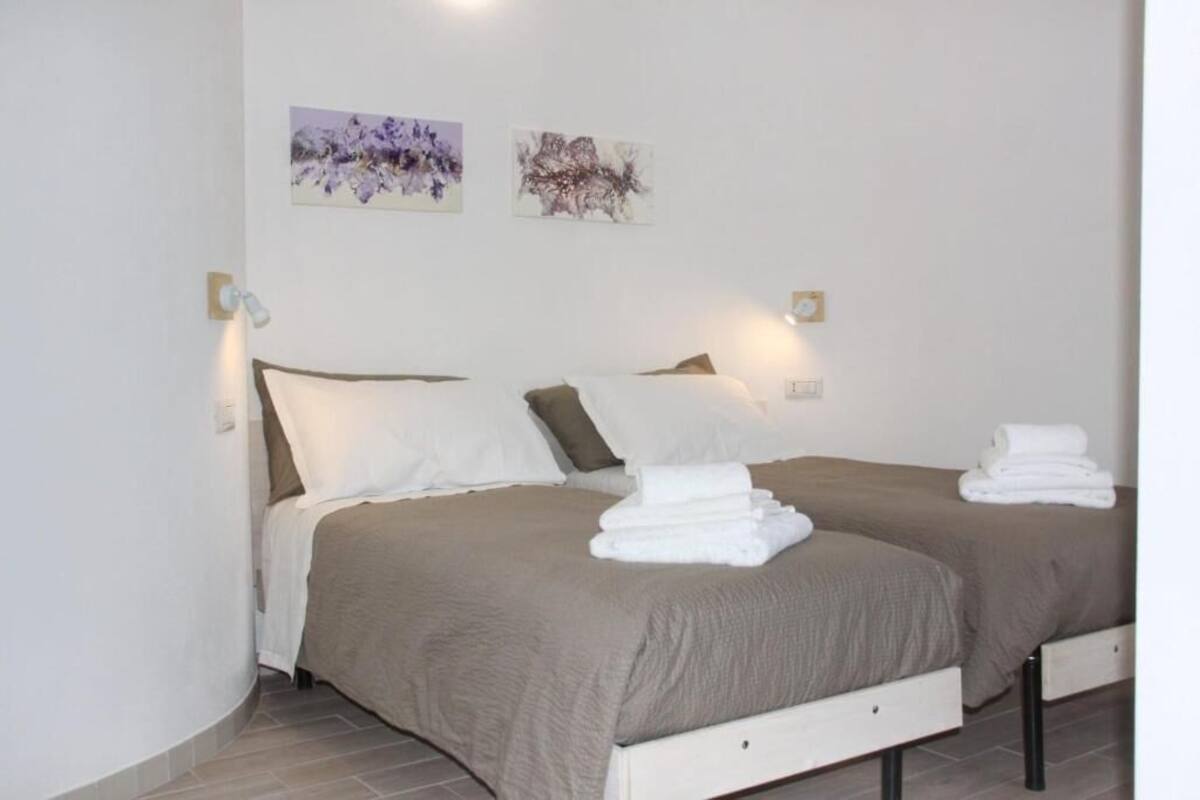 A double bed with a beige bedspread, two pillows, and a folded towel on each bed. There are two wall-mounted light fixtures and two abstract paintings on the wall.