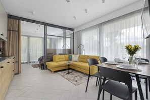Een moderne woonkamer met een gele bank, een eettafel en stoelen. Er is een keukenhoek met kastjes en een aanrecht.