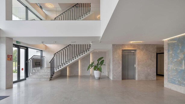 A modern hotel lobby with a glass elevator, a staircase with metal railings, and a potted plant. - Air Conditioned Apartment | Pool | Terrace | Close to Lake and Beach (Mielno)