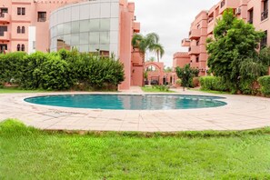 A swimming pool surrounded by a courtyard with a modern building, lush greenery, and a traditional archway. - Gigi Apartment with Pool & Parking (Marrakesh)