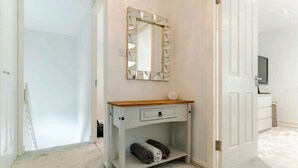 Ein Zimmer mit einer weißen Tür, einem Holzkonsolentisch mit Schublade, einem Spiegel mit geometrischem Rahmen und einem Paar zusammengerollter Handtücher auf dem Boden.