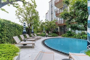 Zona de piscina con sillones de descanso y sombrillas, rodeada de vegetación y un edificio con balcones.