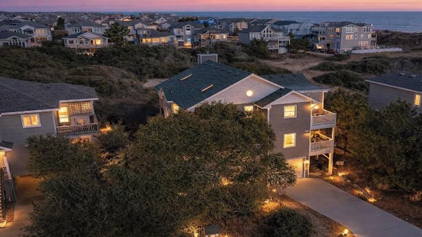 Uma área residencial costeira com casas que têm varandas, um calçadão pavimentado e vista para o oceano ao entardecer.