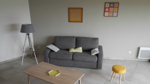 Un salon avec un canapé gris, une table basse en bois, un pouf jaune, une lampe sur pied et deux œuvres d’art murales.