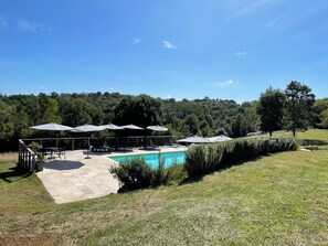 Un espace piscine doté de chaises longues et de parasols, entouré d’une pelouse verdoyante et d’arbres.