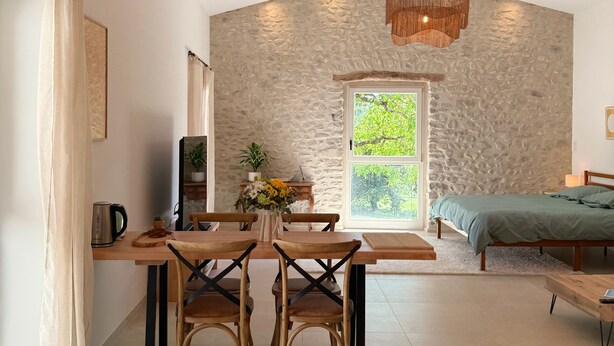 A cozy room with a stone wall, wooden ceiling beams, a dining table with chairs, a bed, and a hanging light fixture. - ⭐​ The Colibris Loft ⭐​ (Mirabel-et-Blacons)