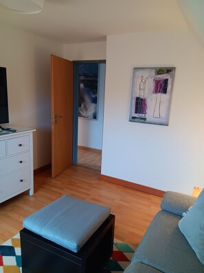 A room with a TV, a sofa, a small ottoman, a door leading to another room, and a wall with artwork. - Vacation rental right on the North Sea in Butjadingen-Tossens (Butjadingen)