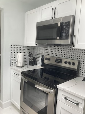 Una cocina con gabinetes blancos, un microondas de acero inoxidable, una estufa negra y un salpicadero de azulejos.