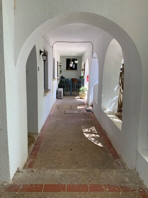 Ein Korridor mit weißen Wänden, einem Ziegelboden und Bögen. Ein Fenster mit hängenden Pflanzen und eine Wand-Klimaanlage sind vorhanden.