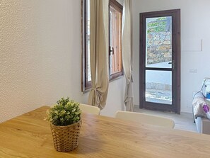 Ein Zimmer mit Holztisch, Topfpflanze, einem Fenster mit Vorhängen und Blick auf eine Mauer vor dem Haus.