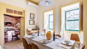 A dining area with a table set for four, a kitchen visible through an open door, and a window with a view of trees. - Quinta das Amendoeiras (Carrazeda de Ansiaes)
