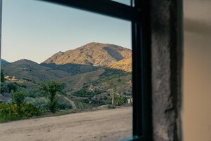 Vista pela janela de uma paisagem montanhosa com uma estrada de terra e vegetação rala.