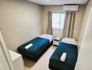 Une petite chambre propre, avec deux lits simples, une table de chevet, une fenêtre avec des rideaux et un appareil de climatisation.