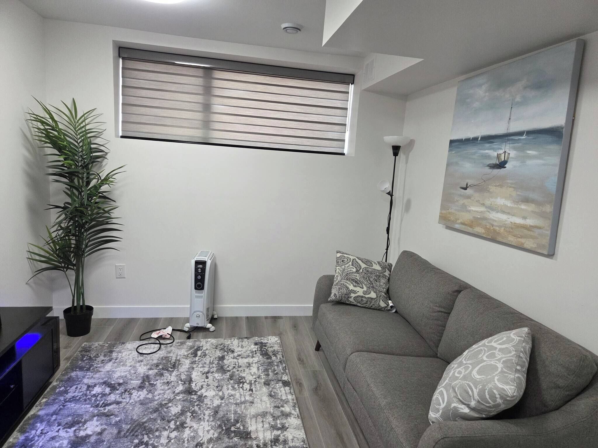 A living room with a gray sofa, a patterned throw pillow, a floor lamp, a large window with blinds, a potted plant, and a painting of a sailboat on the wall.
