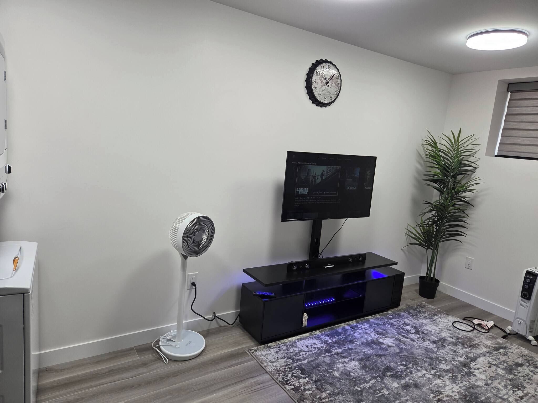 A living room with a flat-screen TV, a standing fan, a clock, a potted plant, and a modern entertainment unit.