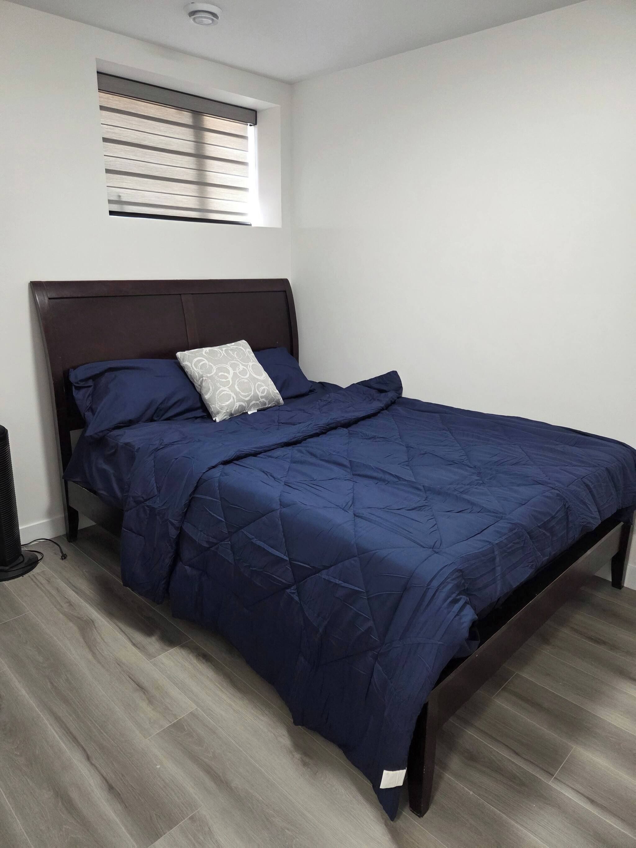 Une chambre à coucher avec un cadre de lit en bois foncé, une couvre-lit matelassée bleue, un oreiller orné d’un motif circulaire et une fenêtre avec des stores horizontaux.