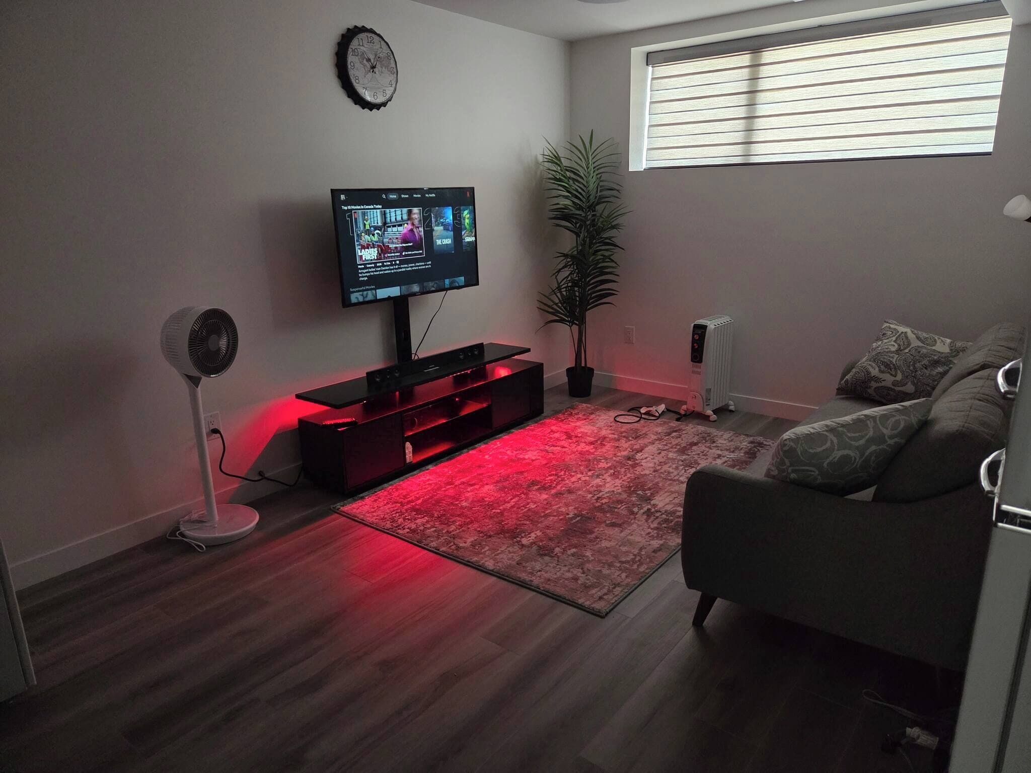 Ein Wohnzimmer mit Flachbildfernseher, Standventilator, Topfpflanze, Heizkörper und einem Fenster mit Jalousien.