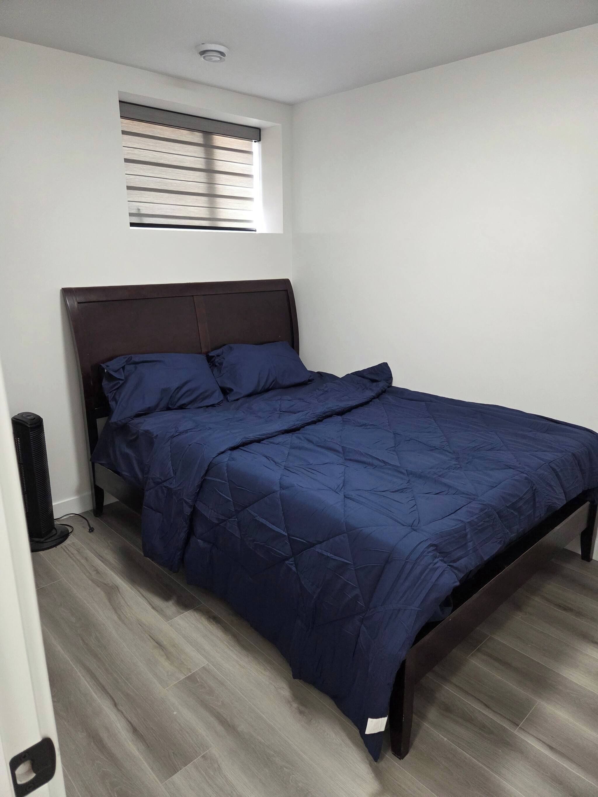 A bedroom with a dark wooden bed frame, a blue quilted bedspread, and a small window with blinds.